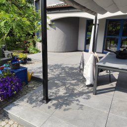 KER BUD - Nowoczesny taras z szarych płyt, zadaszony pergolą. Widoczne kwiaty w donicach i meble ogrodowe. Cień drzew na płytach. Jasne, słoneczne oświetlenie.