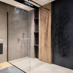 Nowoczesna kabina prysznicowa walk-in z szarymi płytkami, drewnianym panelem i czarnym grzejnikiem dekoracyjnym w kształcie plastra miodu na ciemnej ścianie.