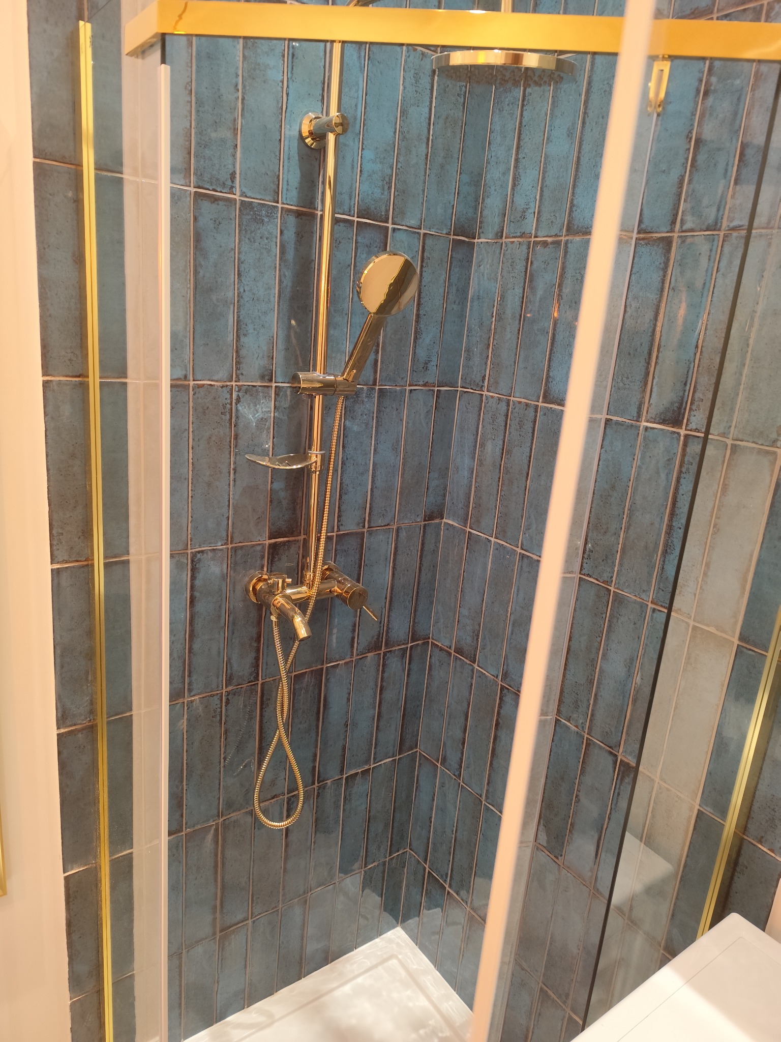 Kabina prysznicowa z niebieskimi, pionowo ułożonymi płytkami i złotą armaturą, widok przez częściowo zamgloną szybę.