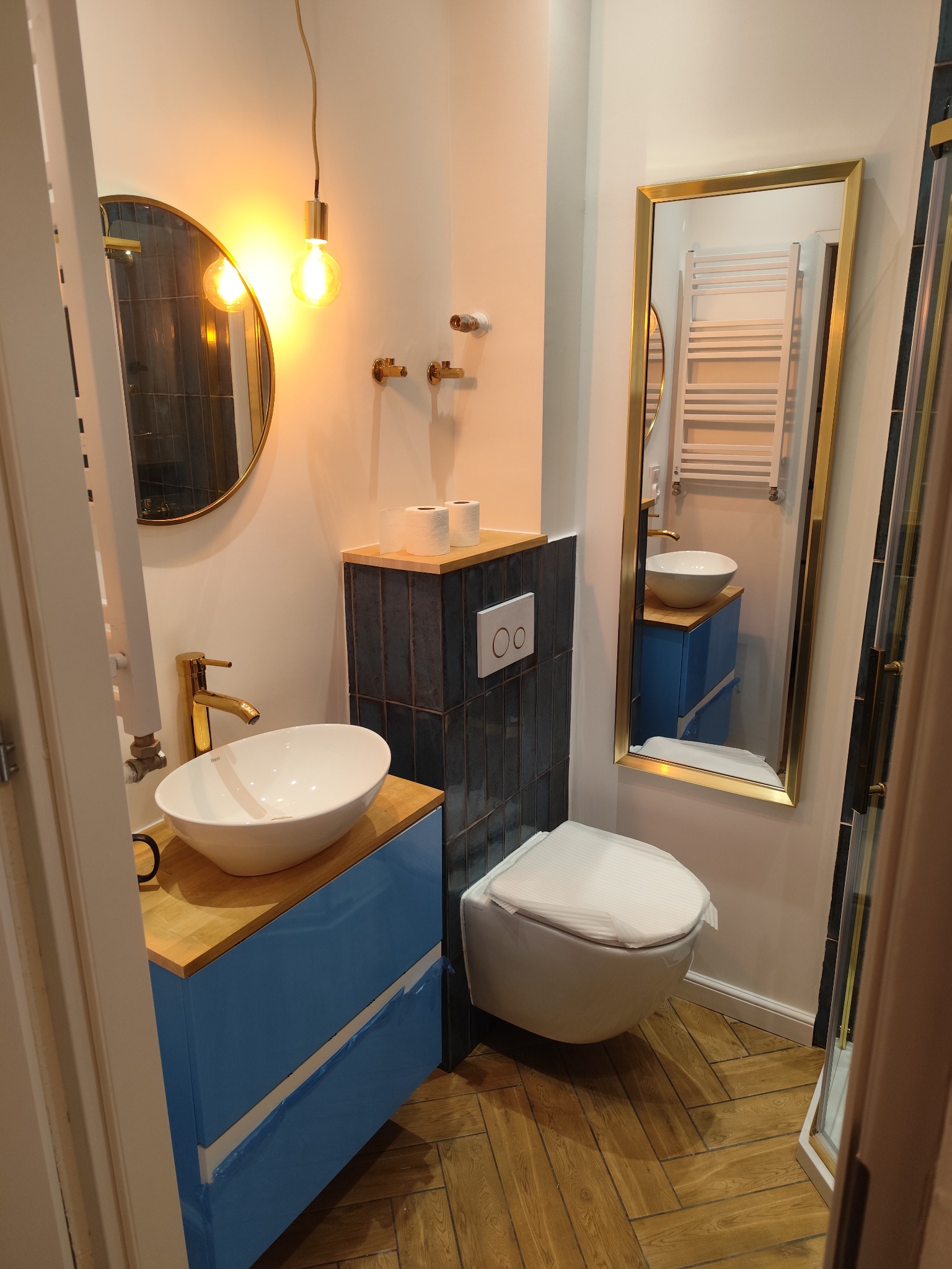 Nowoczesna łazienka z umywalką nablatową, szafką w kolorze błękitu, toaletą wiszącą i lustrem w złotej ramie, widoczna drewniana podłoga ułożona w jodełkę.