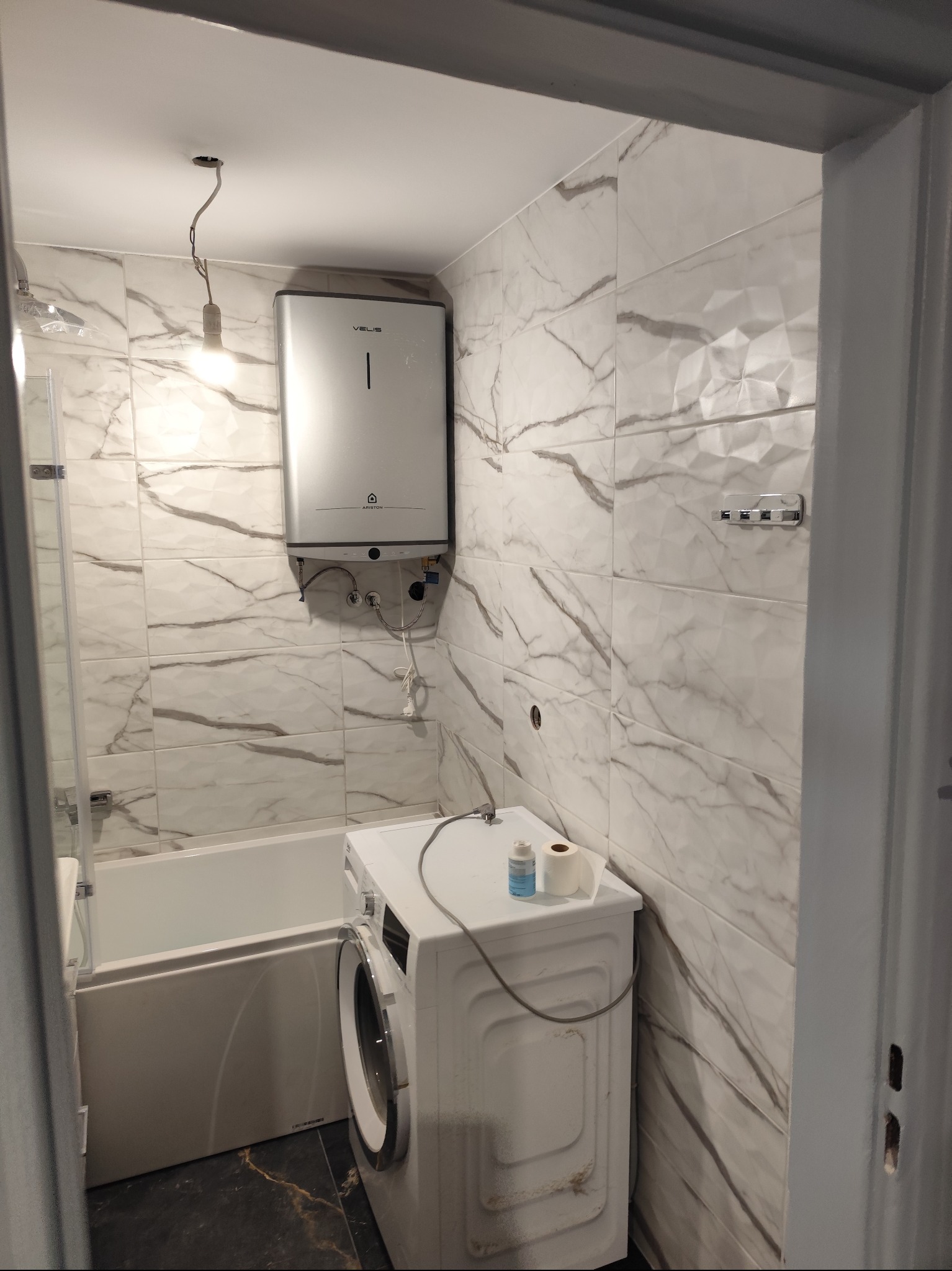 Wykończona łazienka z marmurowymi płytkami, bojlerem elektrycznym i pralką, widok na fragment wanny i czarną podłogę.