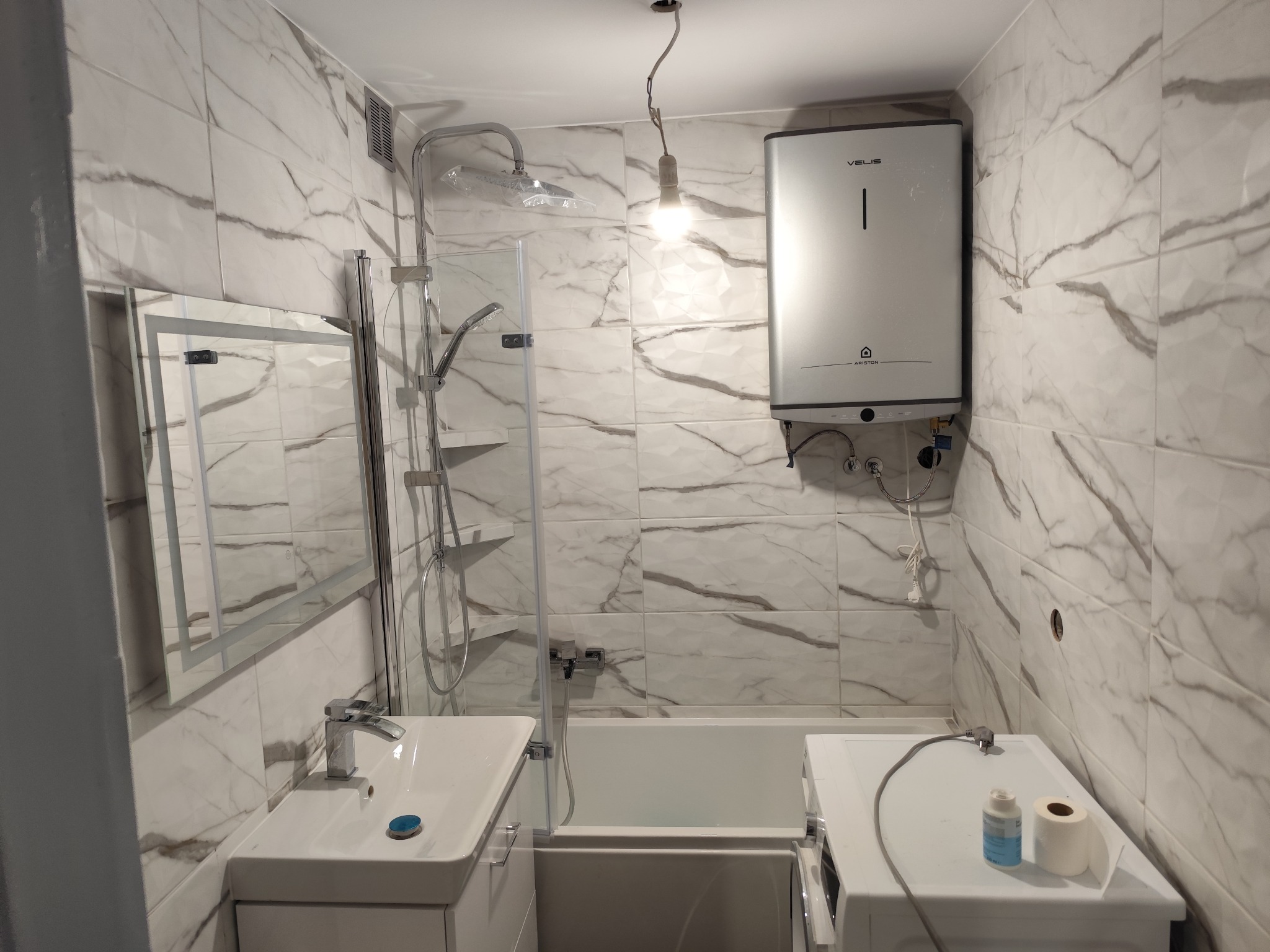Wykończona łazienka z marmurowymi płytkami, kabiną prysznicową, umywalką, wanną, pralką i podgrzewaczem wody Velis Ariston.