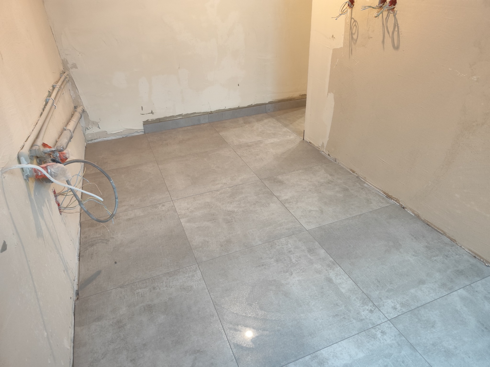 Szare płytki podłogowe ułożone w pomieszczeniu w trakcie remontu, widoczne instalacje na ścianach i ślady po pracach wykończeniowych.
