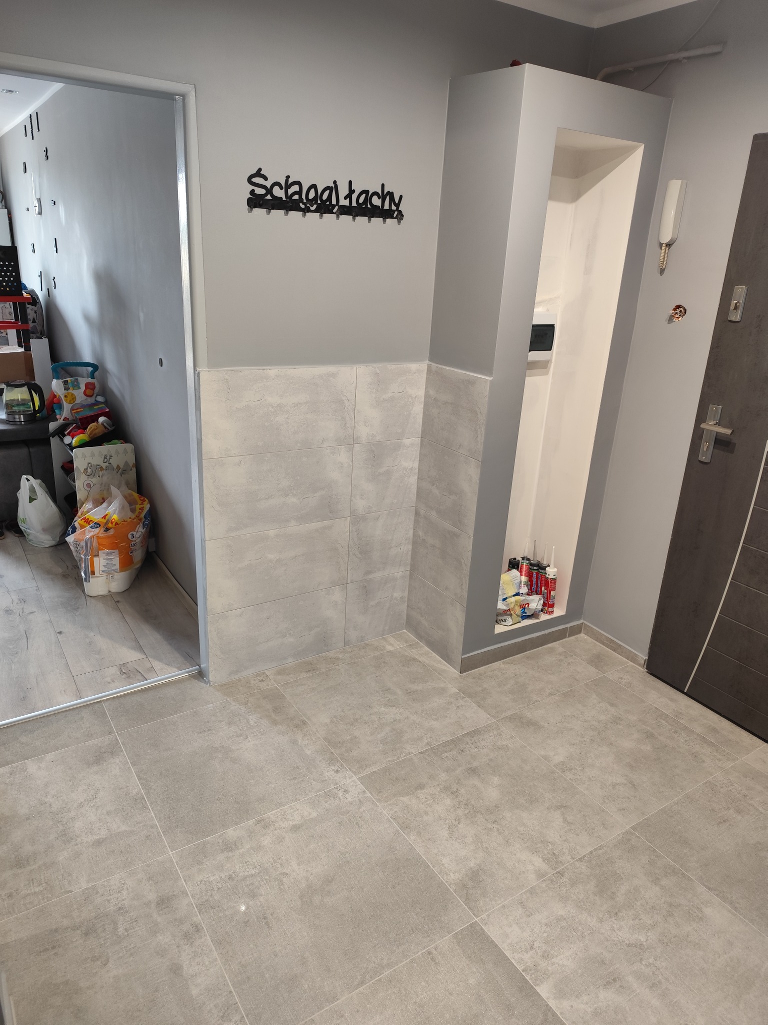 Wykończony korytarz z szarymi płytkami podłogowymi i częściowo na ścianie, widoczny wnęka z materiałami budowlanymi i wieszak na ubrania.