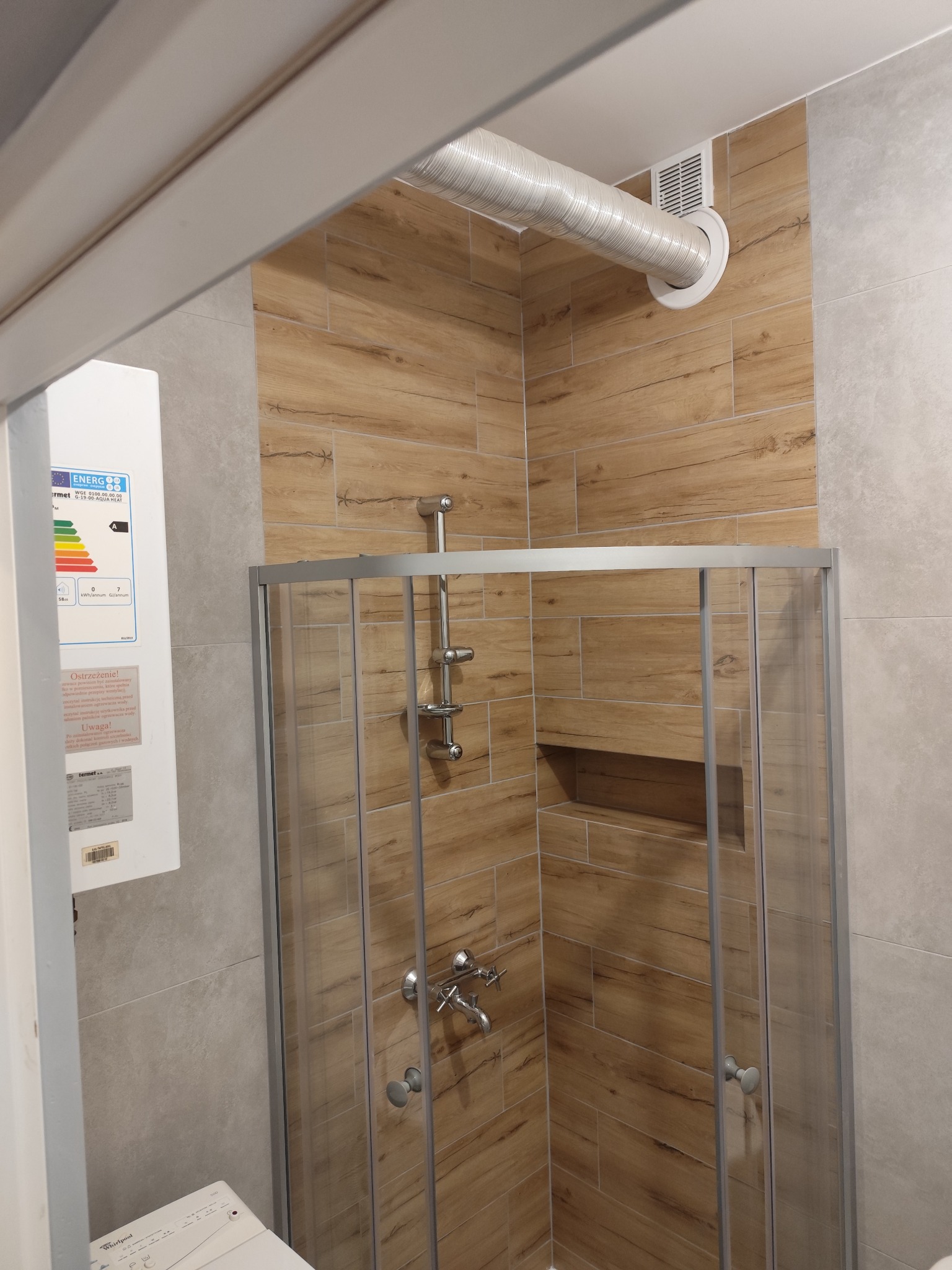 Kabina prysznicowa z drewnopodobnymi płytkami ceramicznymi, widoczny bojler z naklejką energetyczną oraz fragment pralki. W rogu widoczny biały przewód wentylacyjny.