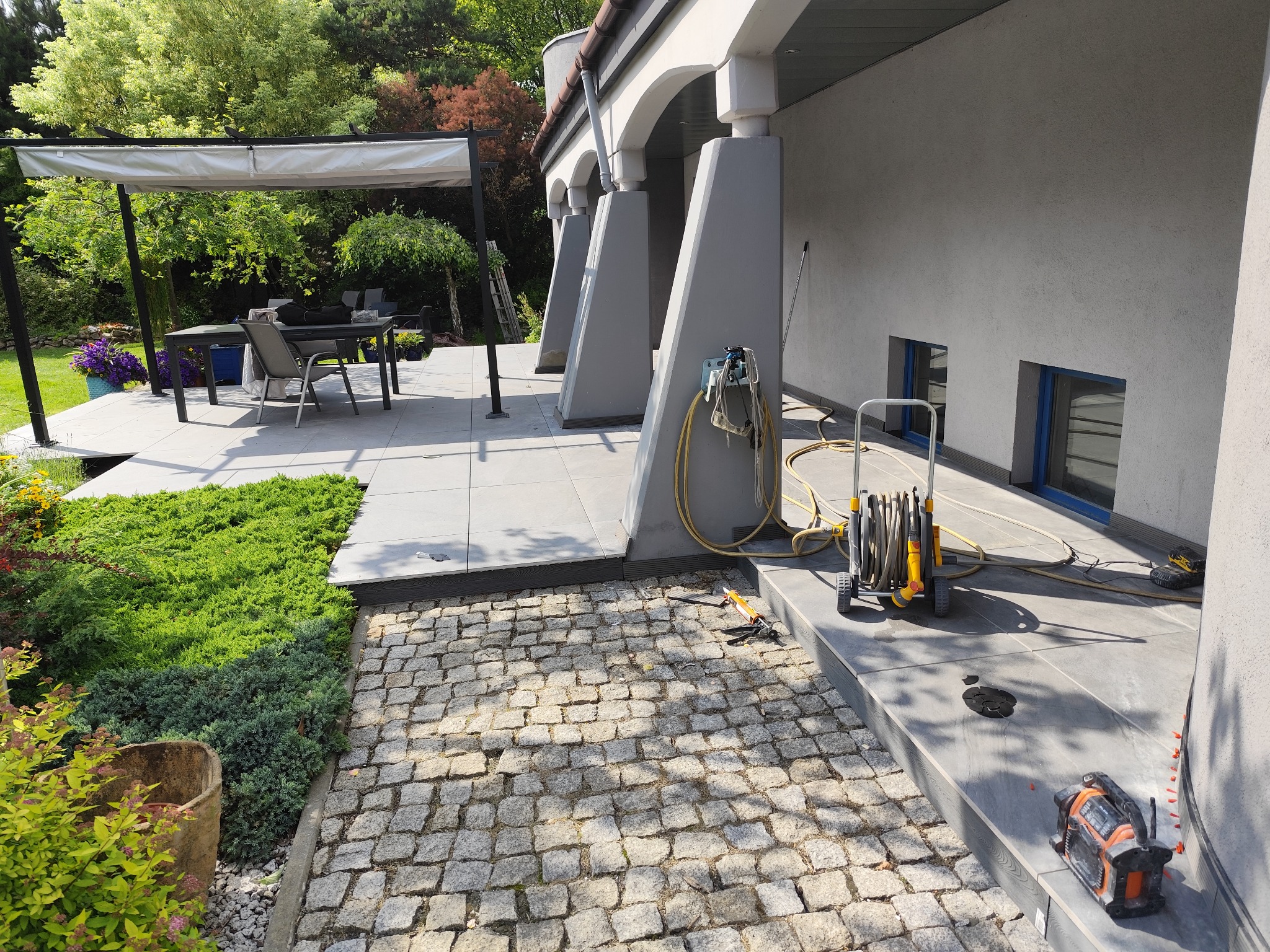 Ułożona kostka brukowa na podjeździe, z widoczną maszyną do czyszczenia ciśnieniowego i narzędziami glazurniczymi. Tło stanowi budynek z tarasem i zielenią.