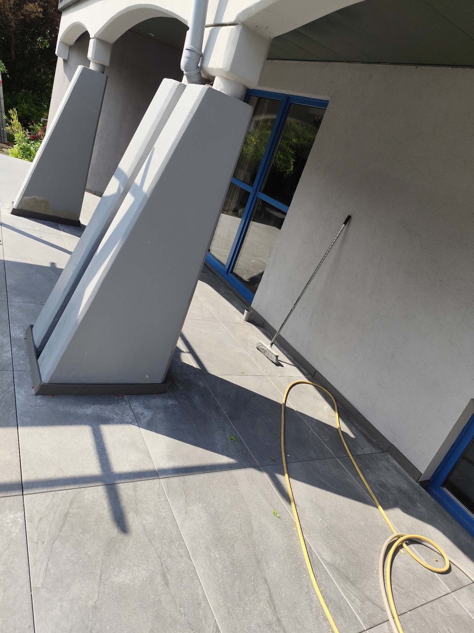 Szare, trapezowe kolumny podtrzymujące zadaszenie, widoczne na tle wyłożonego płytkami tarasu i ściany budynku z niebieskimi ramami okiennymi. W tle zieleń.