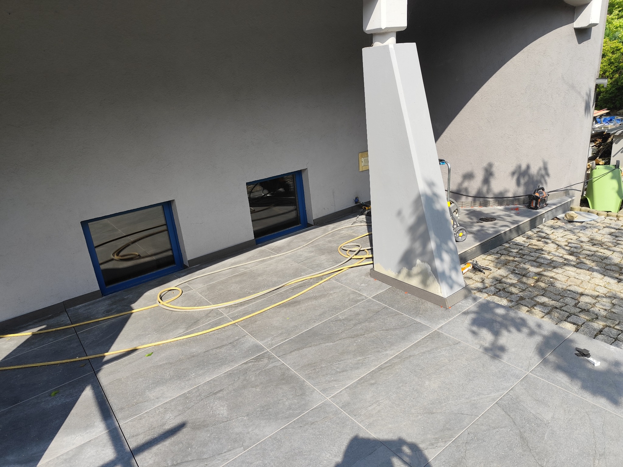 Ułożona szara, matowa terakota na zewnątrz budynku, widoczne narzędzia glazurnicze i fragment myjki ciśnieniowej. Światło słoneczne z cieniem drzewa.