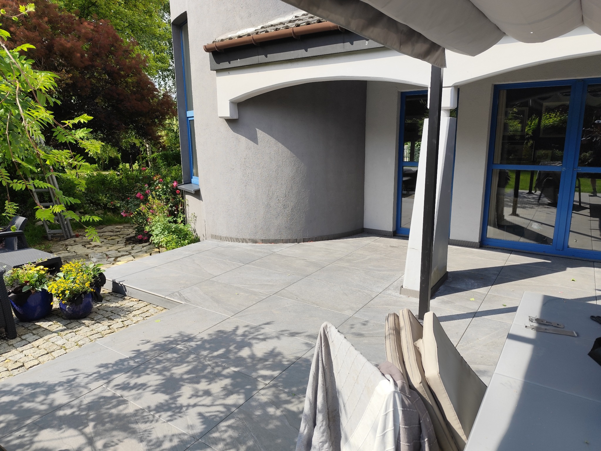 Wyłożony płytkami taras przydomowy z elementami ogrodu, widok z zewnątrz budynku z niebieskimi ramami okiennymi, w tle zieleń i kwiaty.