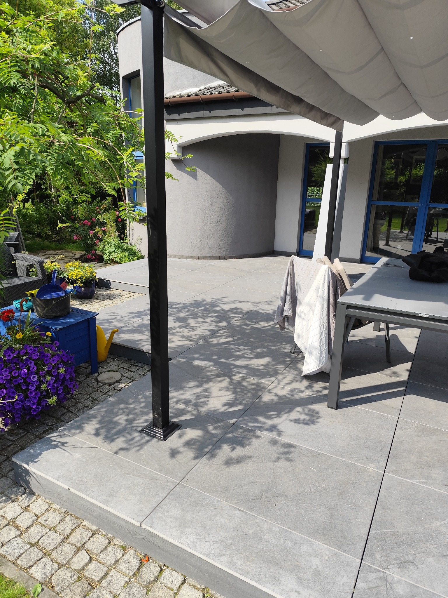 Nowoczesny taras z szarych płyt, zadaszony pergolą. Widoczne kwiaty w donicach i meble ogrodowe. Cień drzew na płytach. Jasne, słoneczne oświetlenie.