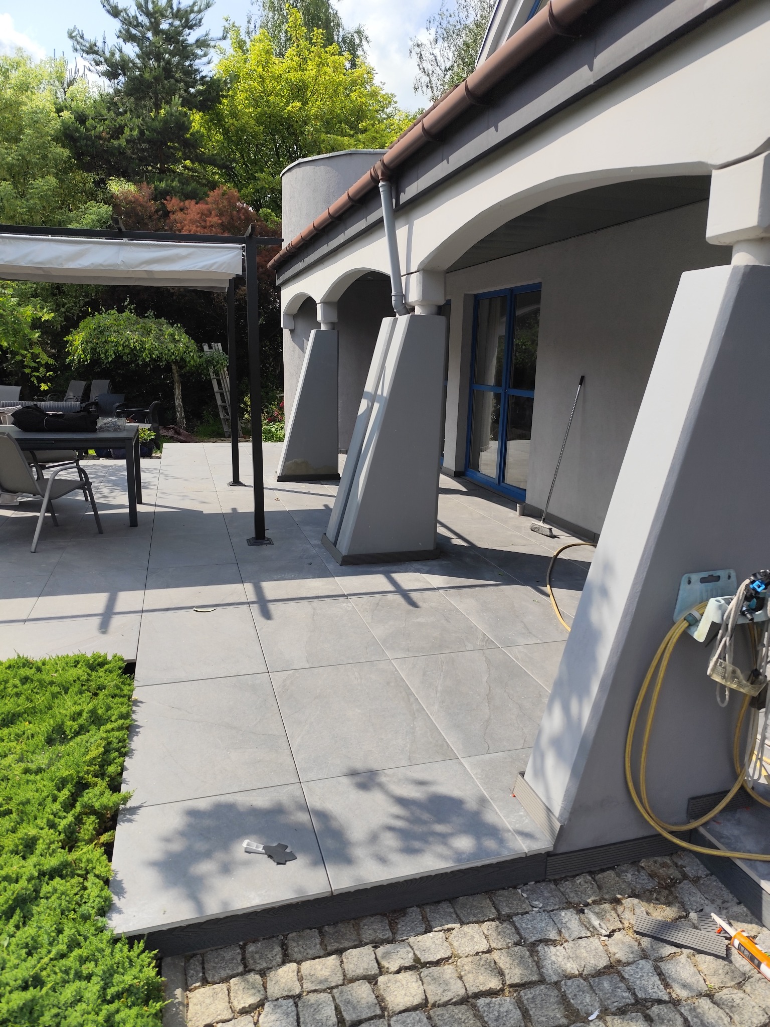 Wyłożony płytkami taras z kolumnami wspierającymi zadaszenie. Widoczne narzędzia i materiały budowlane. Tło stanowi zieleń drzew i krzewów.