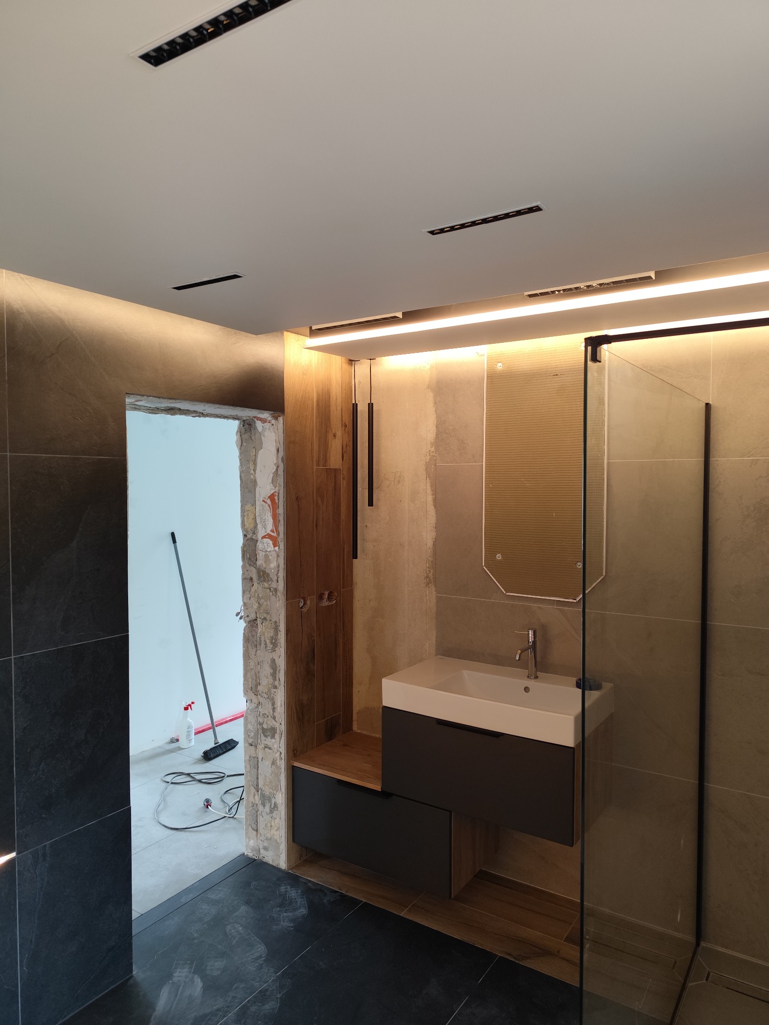 Wykończona łazienka z umywalką w szafce, kabiną prysznicową i oświetleniem LED. Widoczny fragment podłogi i drzwi w trakcie remontu.