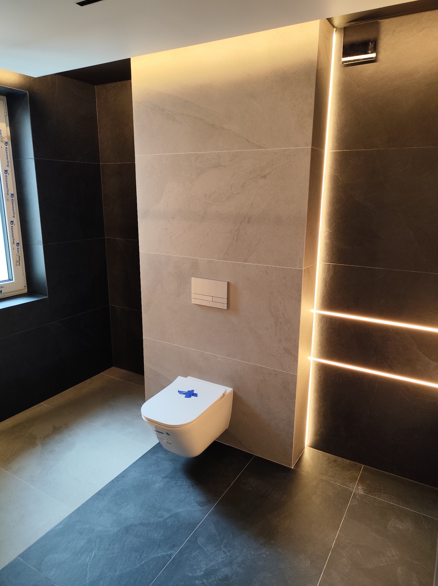 Nowoczesna łazienka z podświetlanymi płytkami ściennymi, podwieszana toaleta z niebieskim zabezpieczeniem. Kontrastujące szare i czarne płytki podłogowe i ścienne.