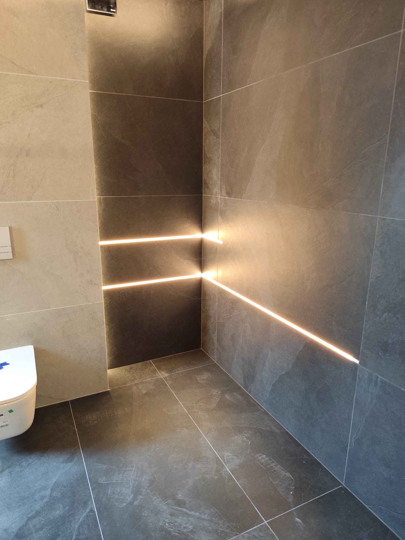 Nowoczesna łazienka z szarymi płytkami i liniowym oświetleniem LED w narożniku. Widoczna toaleta i ślady na podłodze.