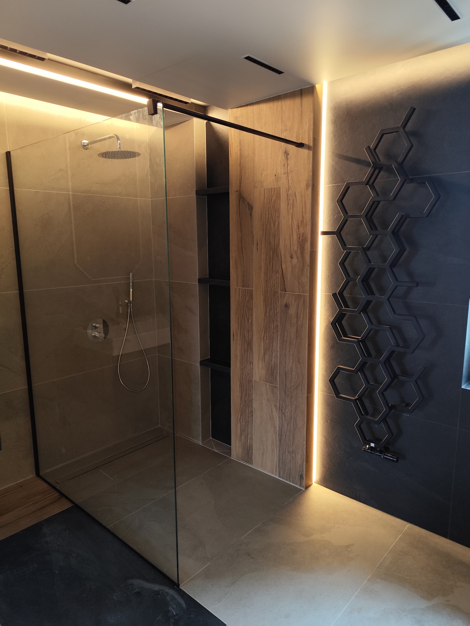 Nowoczesna kabina prysznicowa typu walk-in z deszczownicą, wykończona płytkami w odcieniach beżu i szarości, z dekoracyjnym grzejnikiem w kształcie plastra miodu.