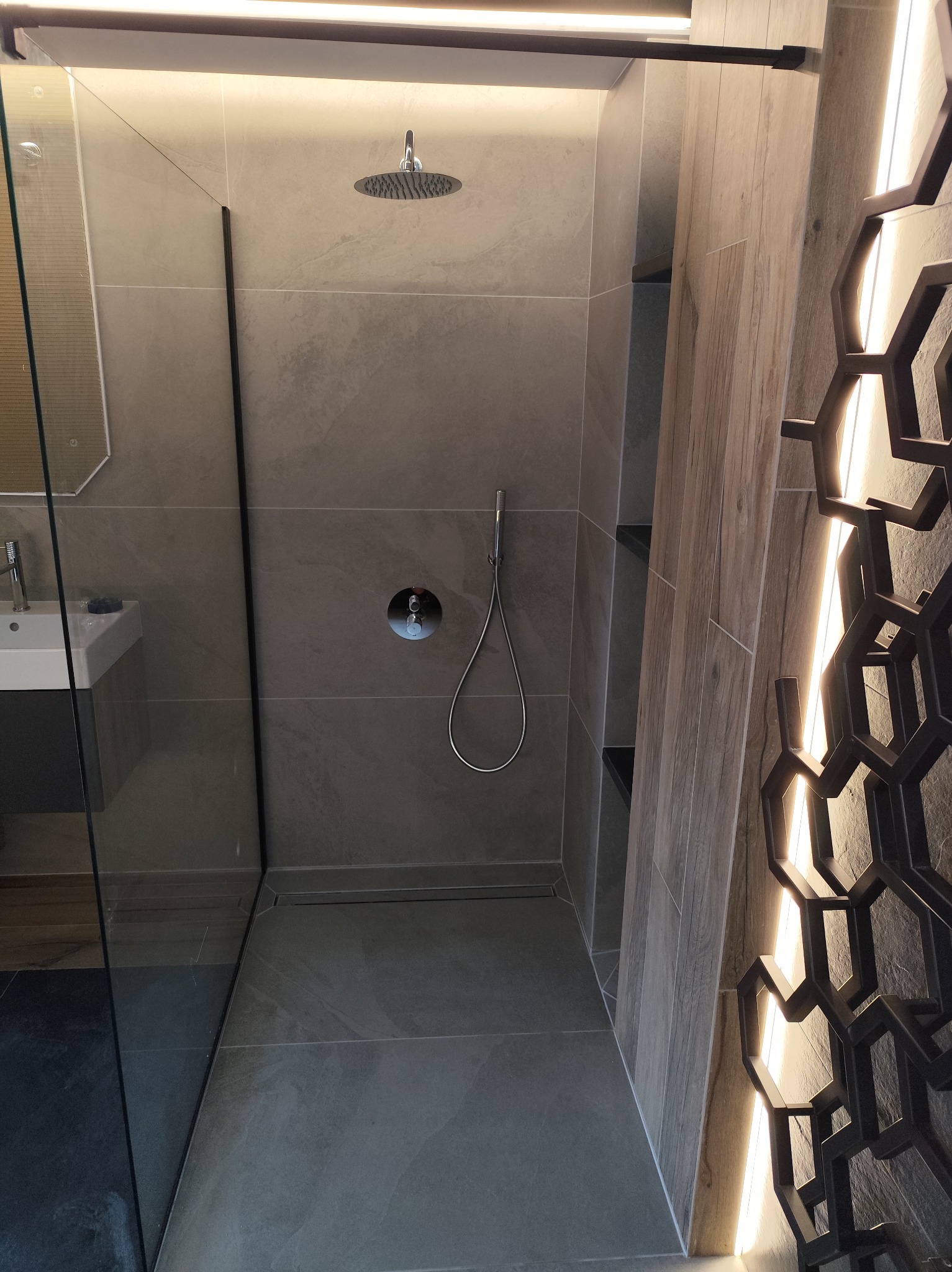 Nowoczesna kabina prysznicowa z szarymi płytkami, deszczownicą i dekoracyjnym oświetleniem w kształcie plastra miodu. Widoczna szklana ściana i umywalka.