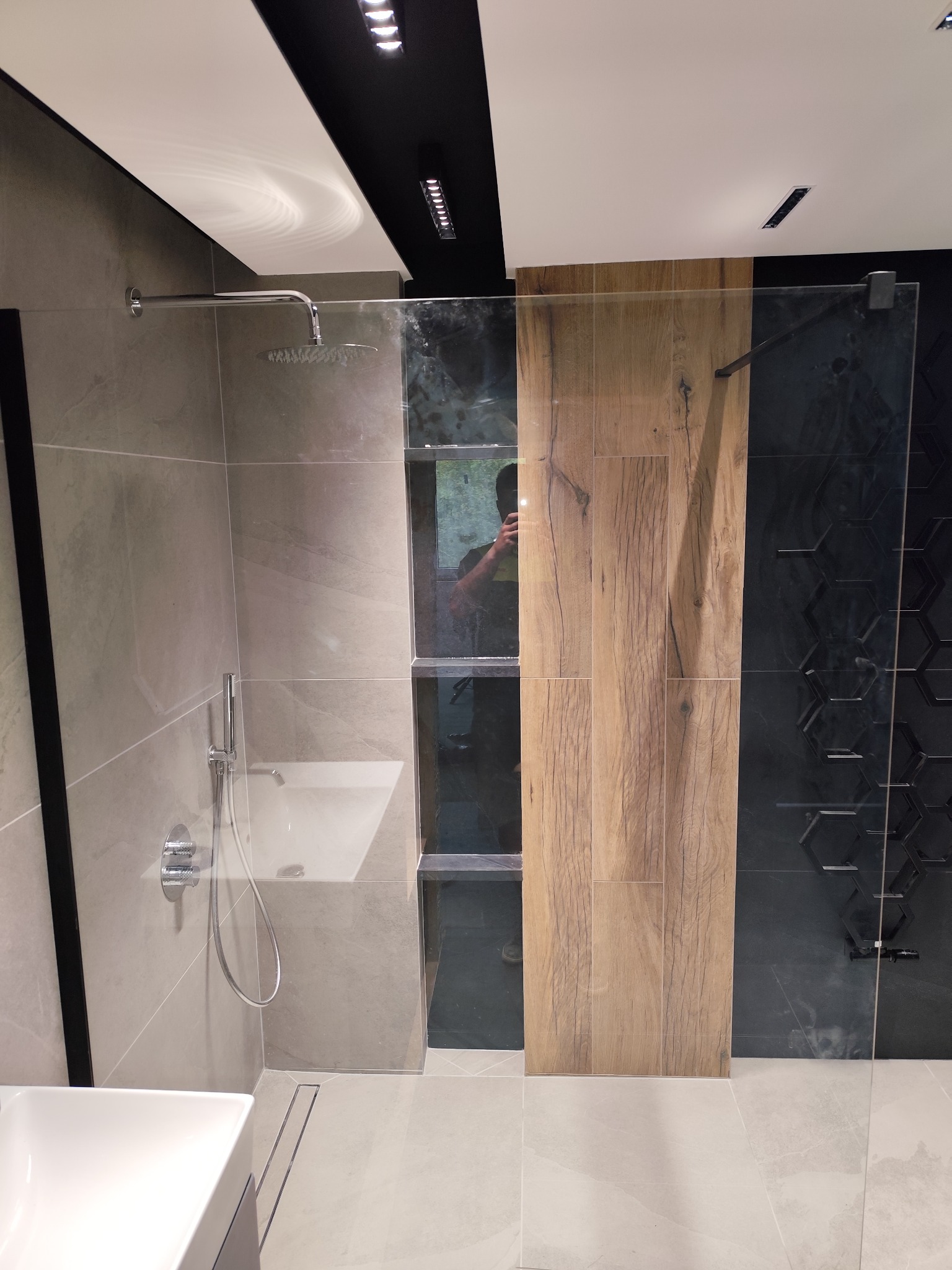 Nowoczesna kabina prysznicowa walk-in z drewnianą ścianką i szarymi płytkami. Widoczna armatura, odpływ liniowy i oświetlenie punktowe.