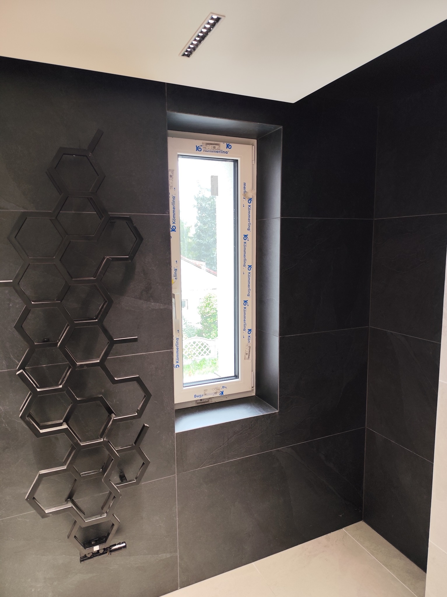 Ciemne płytki w łazience z dekoracyjnym grzejnikiem w kształcie plastra miodu i oknem z widokiem na zieleń. Nowoczesne wykończenie.