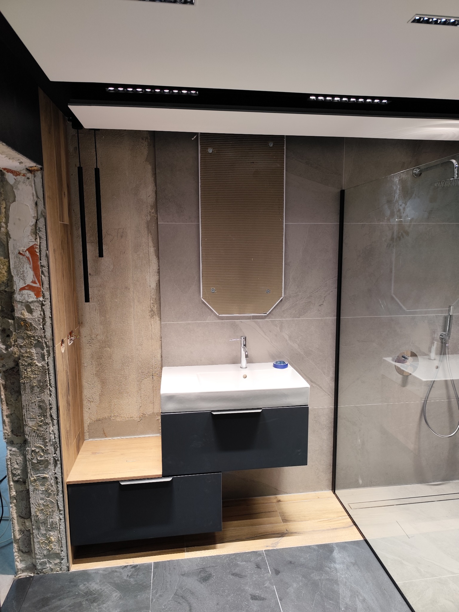 Nowoczesna łazienka z umywalką nablatową, lustrem i kabiną prysznicową typu walk-in. Widoczne szare płytki imitujące kamień i drewniane akcenty. Surowy stan ściany obok.