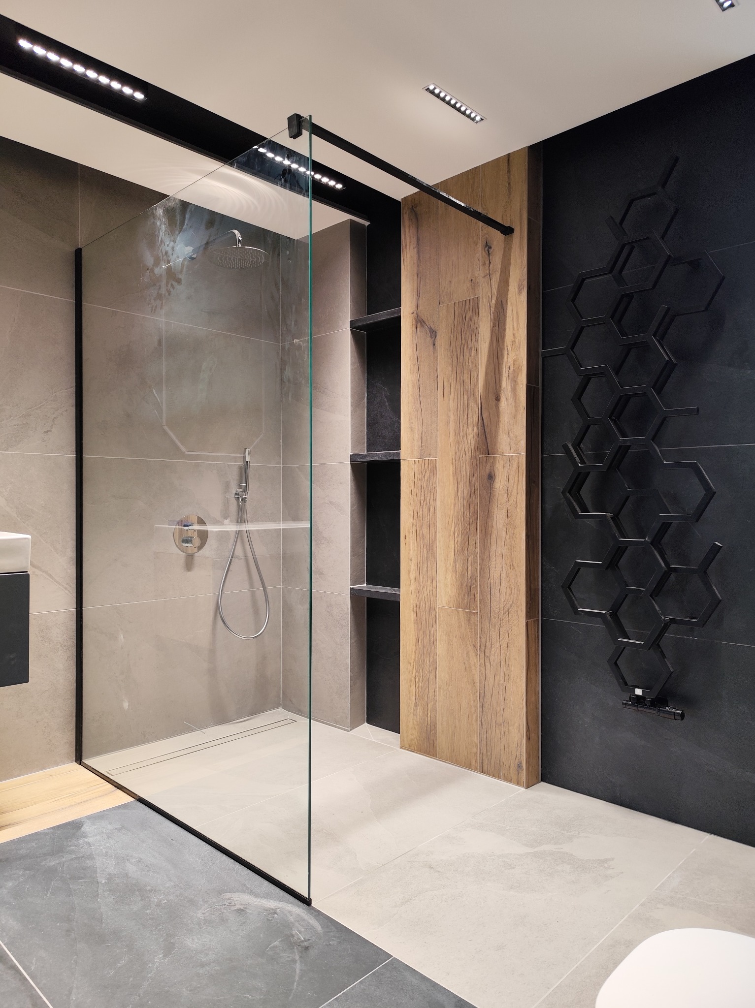 Nowoczesna kabina prysznicowa walk-in z szarymi płytkami, drewnianym panelem i czarnym grzejnikiem dekoracyjnym w kształcie plastra miodu na ciemnej ścianie.