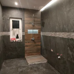 Nowoczesna kabina prysznicowa z ciemnymi, marmurkowymi płytkami i drewnopodobną ścianą w Gorzowie Wielkopolskim; widoczne elementy instalacji i okno z firanką.