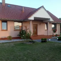 Dom jednorodzinny z beżową elewacją, brązowymi oknami i czerwoną dachówką, widok z ogrodu, z elementami dekoracyjnymi takimi jak róże i lampy kuliste.