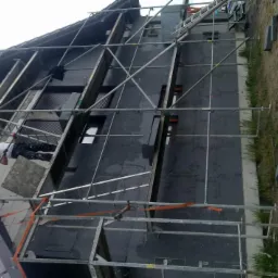 Pracownik na rusztowaniu montuje izolację termiczną na elewacji domu. Widoczne czarne płyty izolacyjne i metalowe rusztowanie. Ujęcie z dołu, lekko pod kątem.