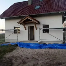 Dom jednorodzinny z białą elewacją w trakcie prac wykończeniowych, widoczne rusztowania i plandeka ochronna, dach z czerwonej dachówki.