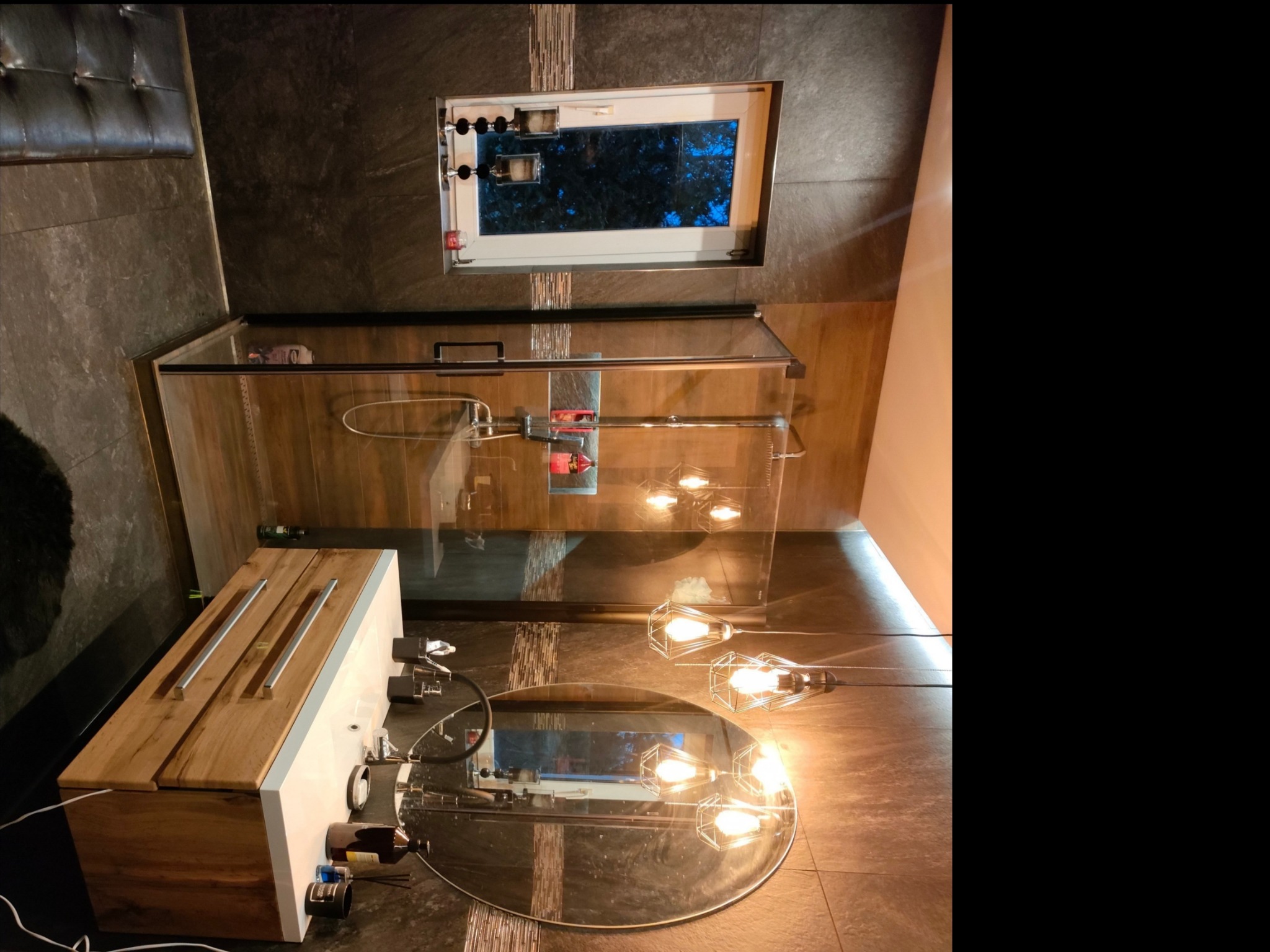 Nowoczesna łazienka z kabiną prysznicową ze szklanymi drzwiami, umywalką nablatową z drewnianym blatem, okrągłym lustrem podświetlanym przez designerskie lampy druciane i oknem z widokiem na zewnątrz.