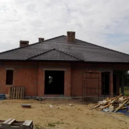 Budowa domu murowanego w stanie surowym otwartym z ciemną dachówką, widoczne rusztowanie i składowane materiały budowlane na placu budowy.