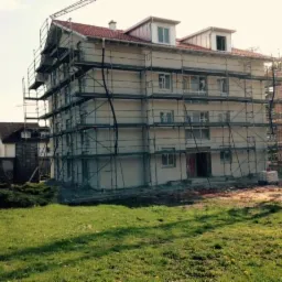 Wielopiętrowy budynek w trakcie prac elewacyjnych, otoczony rusztowaniami. Widoczne fragmenty elewacji pokryte tynkiem. W tle zieleń i inne budynki.