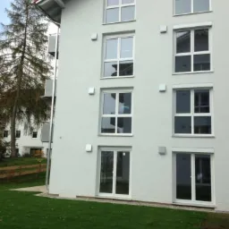 Elewacja budynku mieszkalnego po remoncie, z jasną fasadą, białymi oknami i roletami na niektórych oknach. Widoczne trawnik i drzewo.