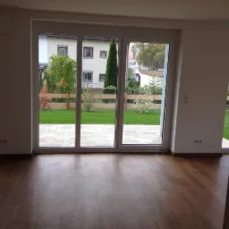 Nowe panele podłogowe w jasnym pokoju z dużym oknem tarasowym i widokiem na ogród. Ciemne panele i białe ściany tworzą kontrast.