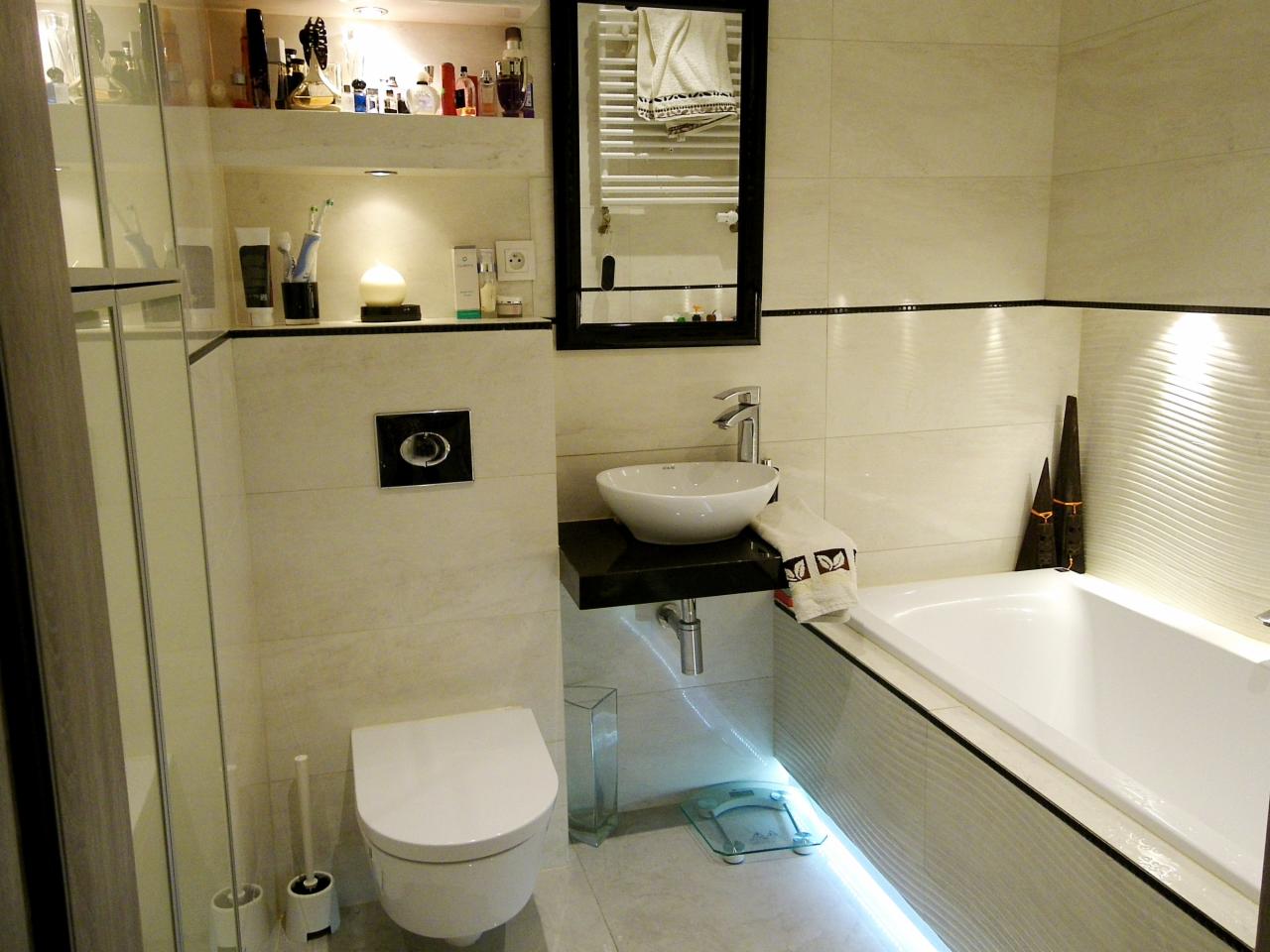 Elegancka łazienka z beżowymi płytkami, podświetleniem LED pod wanną, okrągłą umywalką i czarnymi akcentami. Lustro w czarnej ramie i dekoracje na półce.