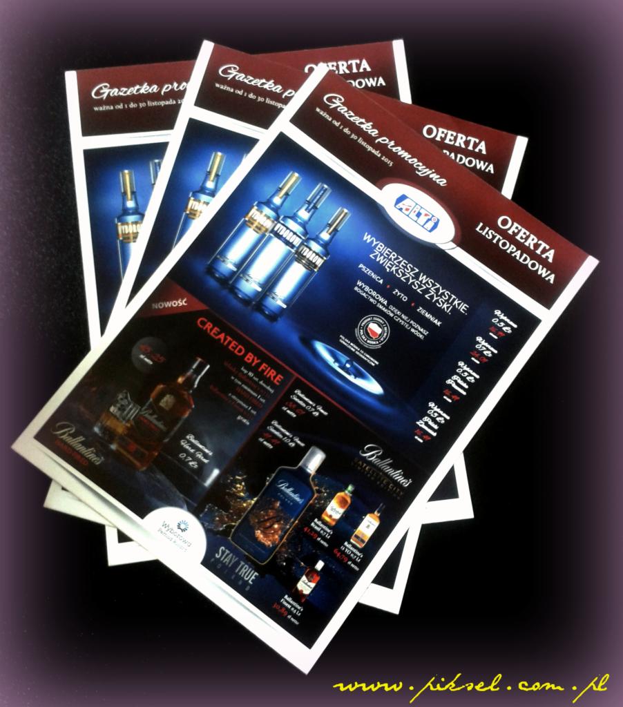 Stos ulotek reklamowych z ofertą promocyjną alkoholi, na ciemnym tle. Widoczne różne marki i ceny. Ulotki z napisem 'Gazetka promocyjna'.