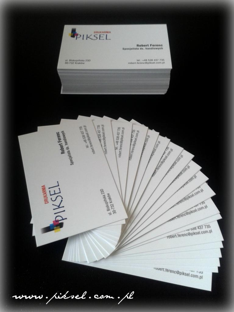 Stos wizytówek firmy Piskel ułożonych wachlarzowo i w stos. Wizytówki z danymi kontaktowymi Roberta Ferenca, specjalisty ds. handlowych, na czarnym tle.