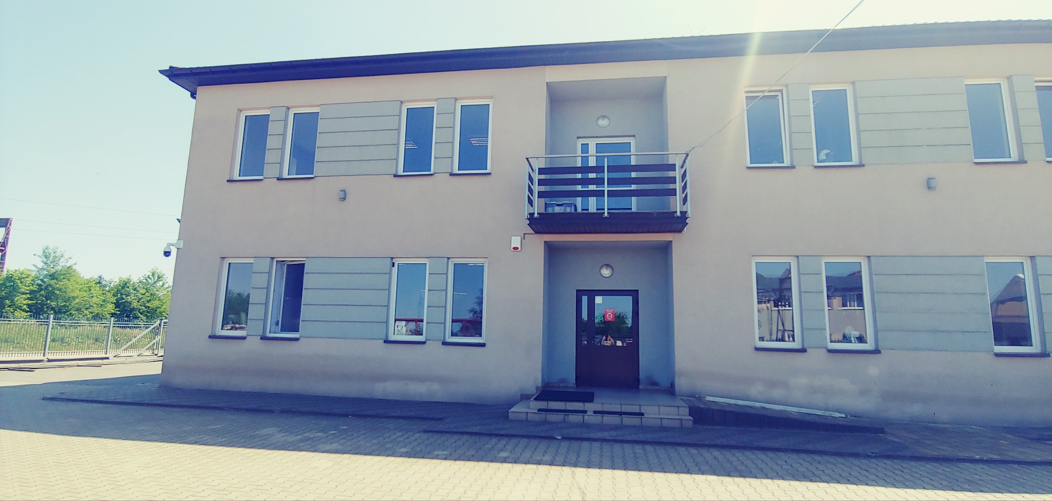 Dwupiętrowy budynek biurowy z balkonem i licznymi oknami, widok z zewnątrz w słoneczny dzień.