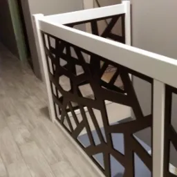 Biała balustrada z dekoracyjnym, geometrycznym wzorem w kolorze ciemnego brązu, zamontowana wewnątrz budynku, widok z góry na drewnianą podłogę.