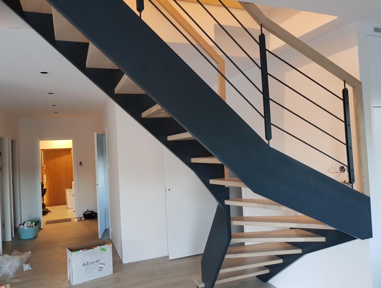 Nowoczesne schody z czarną konstrukcją metalową, jasnymi drewnianymi stopniami i minimalistyczną balustradą z czarnych prętów, widoczne z boku w jasnym wnętrzu z kartonem na podłodze.