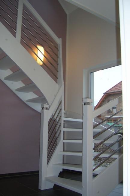 Białe schody z metalowymi balustradami, oświetlone kinkietem, widoczne z dołu, z fragmentem okna i budynku w tle.