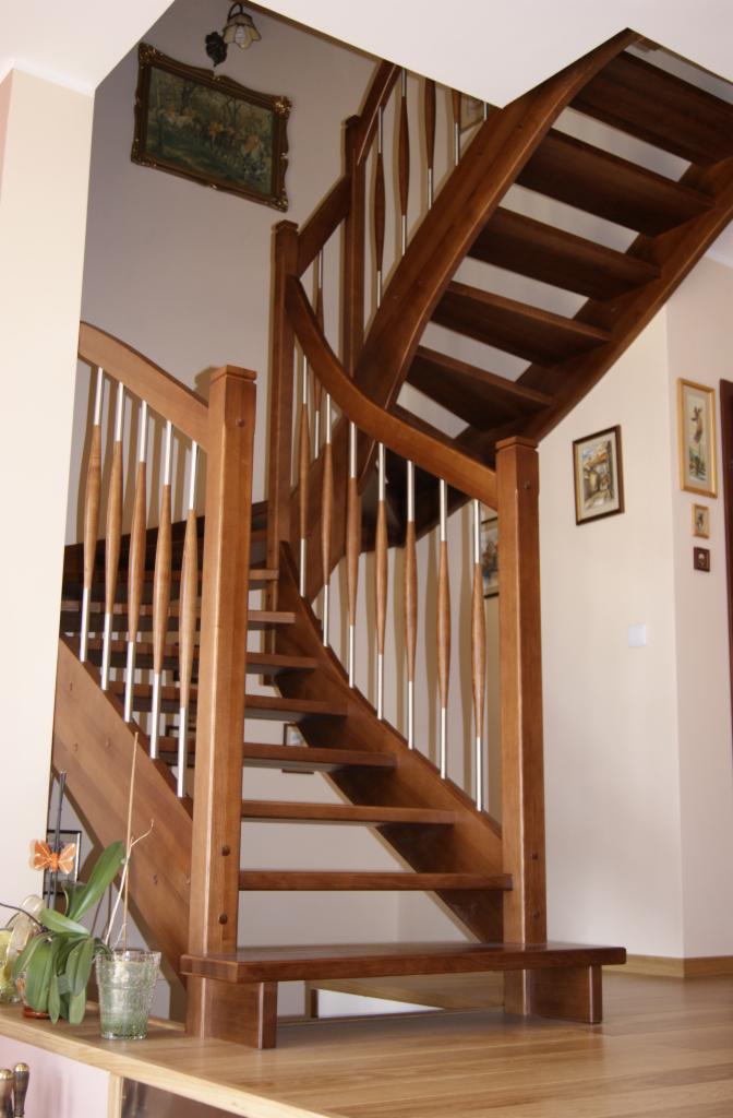 Zakręcone schody z drewna o ciepłym odcieniu, z metalowymi, pionowymi elementami balustrady, widziane z dołu, z kwiatem w doniczce w lewym dolnym rogu.