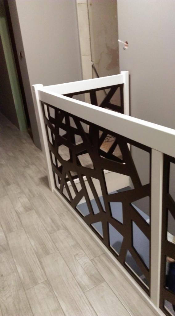 Biała balustrada z dekoracyjnym, geometrycznym wzorem w kolorze ciemnego brązu, zamontowana wewnątrz budynku, widok z góry na drewnianą podłogę.