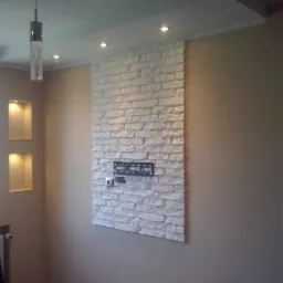 Beżowe wnętrze z dekoracyjną ścianą z białej cegły, na której zamontowano uchwyt na telewizor. Wnęki z oświetleniem i wisząca lampa dodają charakteru.