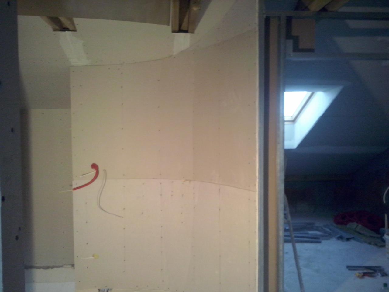 Ściana z płyt karton-gips z widocznymi przewodami elektrycznymi, fragment poddasza z oknem dachowym w tle, surowy stan wykończenia.