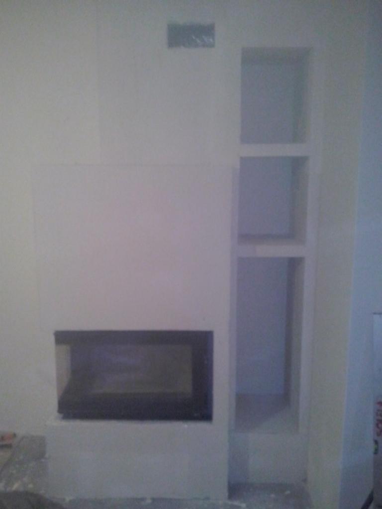 Ściana z wnęką i kominkiem o geometrycznym kształcie, wykończona gładzią, gotowa do malowania. Widoczne detale wykończeniowe.