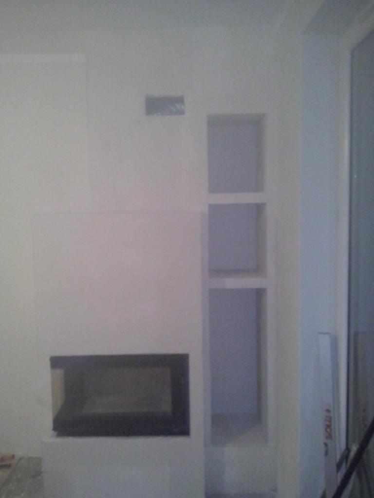 Biała ściana z wnęką na kominek z czarną obudową i trzema półkami w pionie. Widoczne niedoskonałości w wykończeniu.