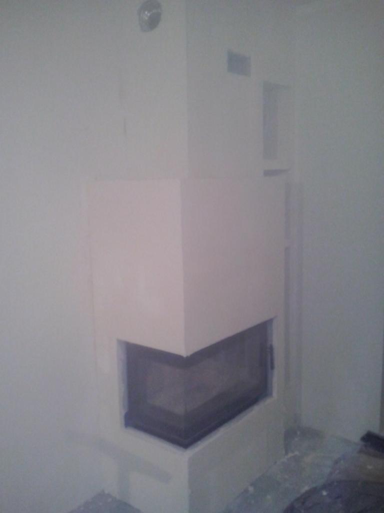 Kanciasty kominek narożny z czarną ramą i jasną obudową, umieszczony w rogu pomieszczenia. Ściany są białe, widoczny fragment podłogi.