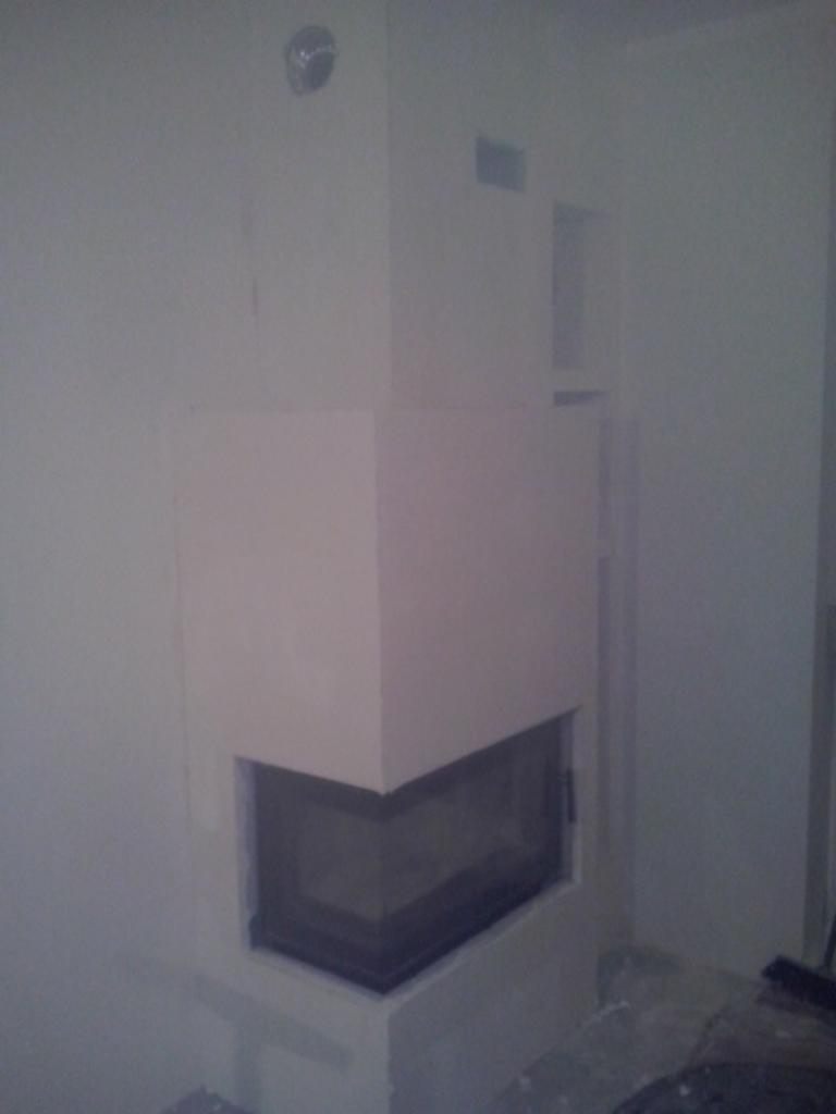 Narożny, surowy kominek obudowany płytami gipsowymi, widoczny w trakcie prac wykończeniowych. Szary odcień ścian i podłogi, otwór wentylacyjny w górnej części.