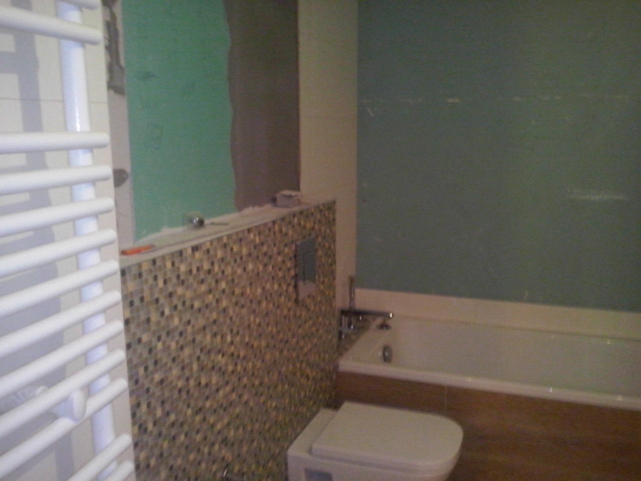 Remont łazienki: częściowo wykończona ściana z mozaiką, fragment zielonej ściany przed położeniem płytek, biały grzejnik drabinkowy i fragment wanny.