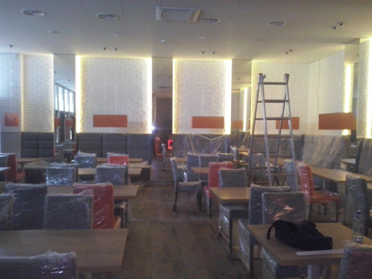 Wnętrze restauracji w trakcie wykańczania: krzesła i stoły zabezpieczone folią, drabina, oświetlenie LED podkreślające ceglane kolumny. Etap prac wykończeniowych.